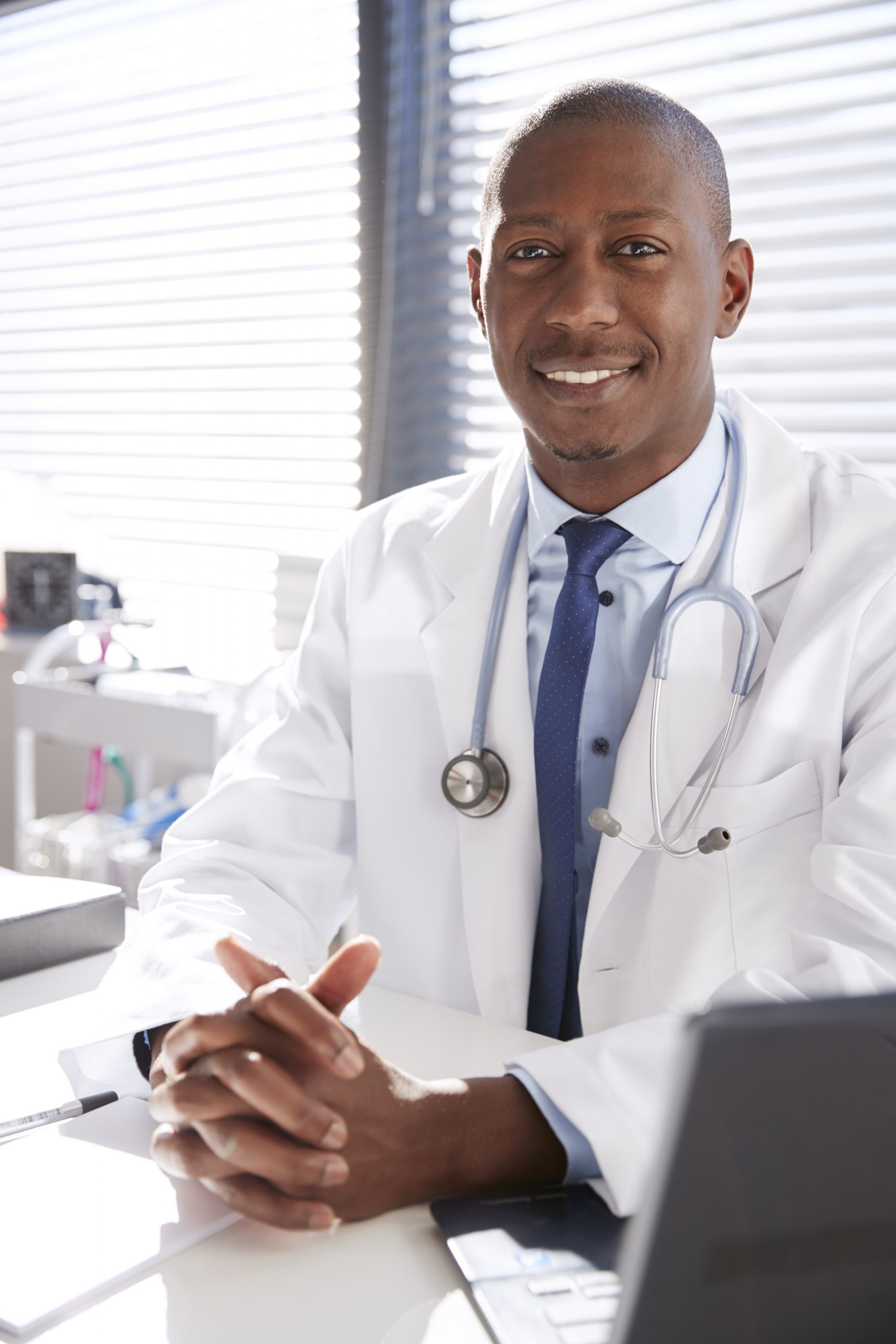 Portrait Of Smiling Male Doctor Wearing White Coat With Stethoscope Sitting Behind Desk In Office
